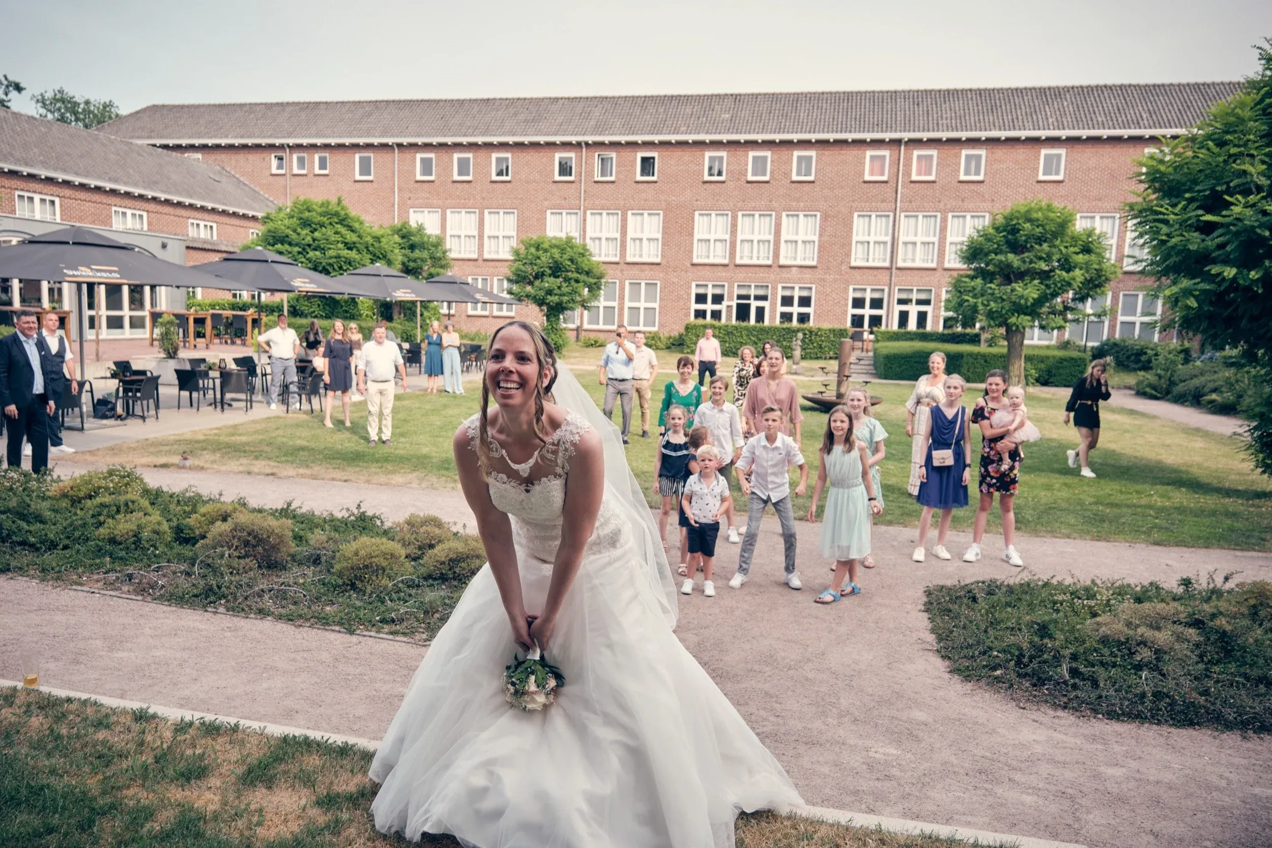 Bruidsmeisje in witte trouwjurk met bloemenboeket, lachend, tijdens bruiloft, met gasten op de achtergrond in tuin.