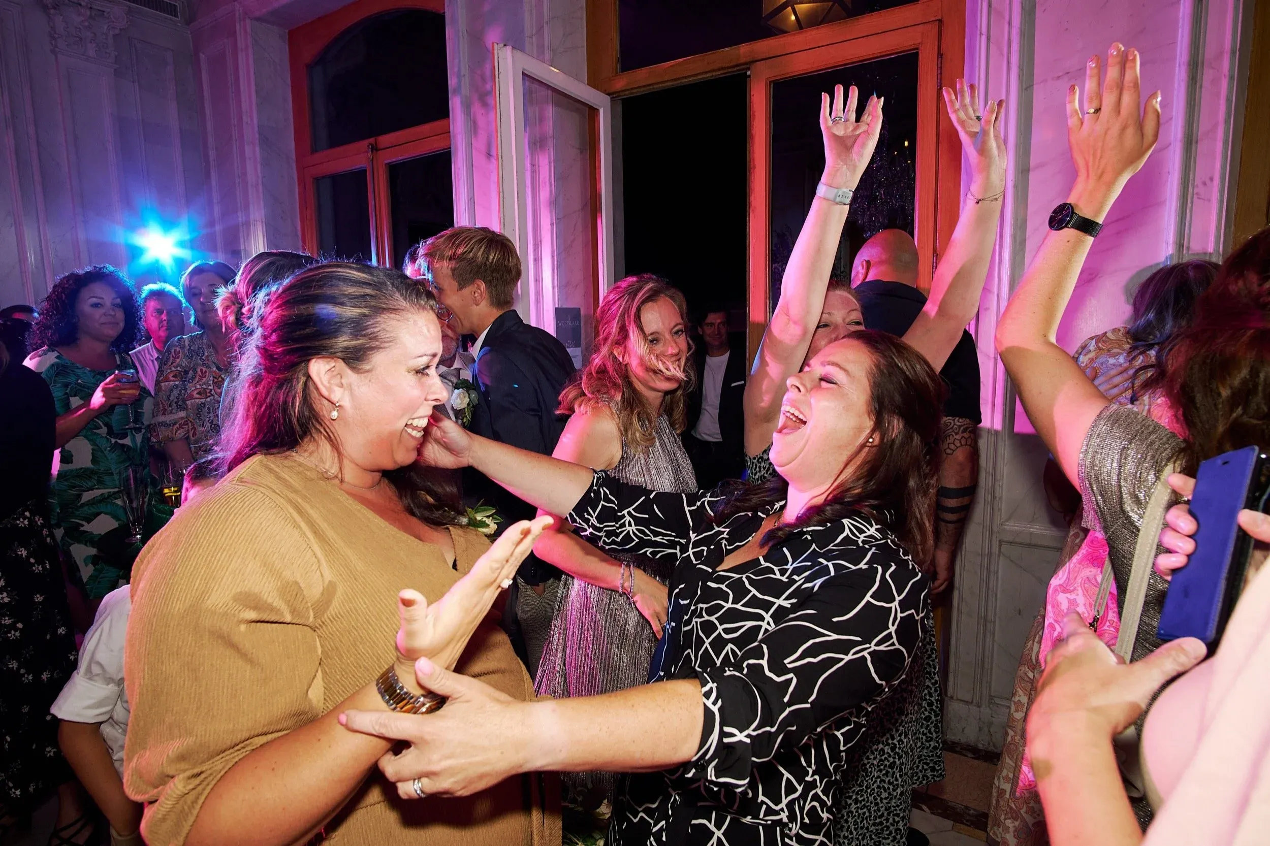 Mensen dansen en lachen op een feest, met kleurrijk licht en open raam in de achtergrond.