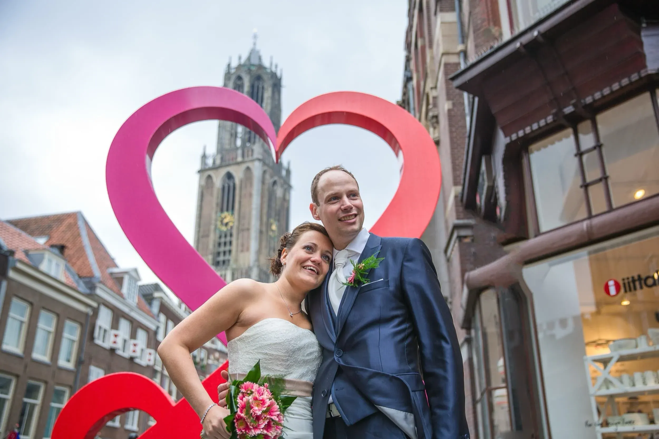 Een gelukkig bruidspaar poseert buiten met een grote roze en rode hartstructuur op de achtergrond, in een straat met historische gebouwen en een gotische kerk.