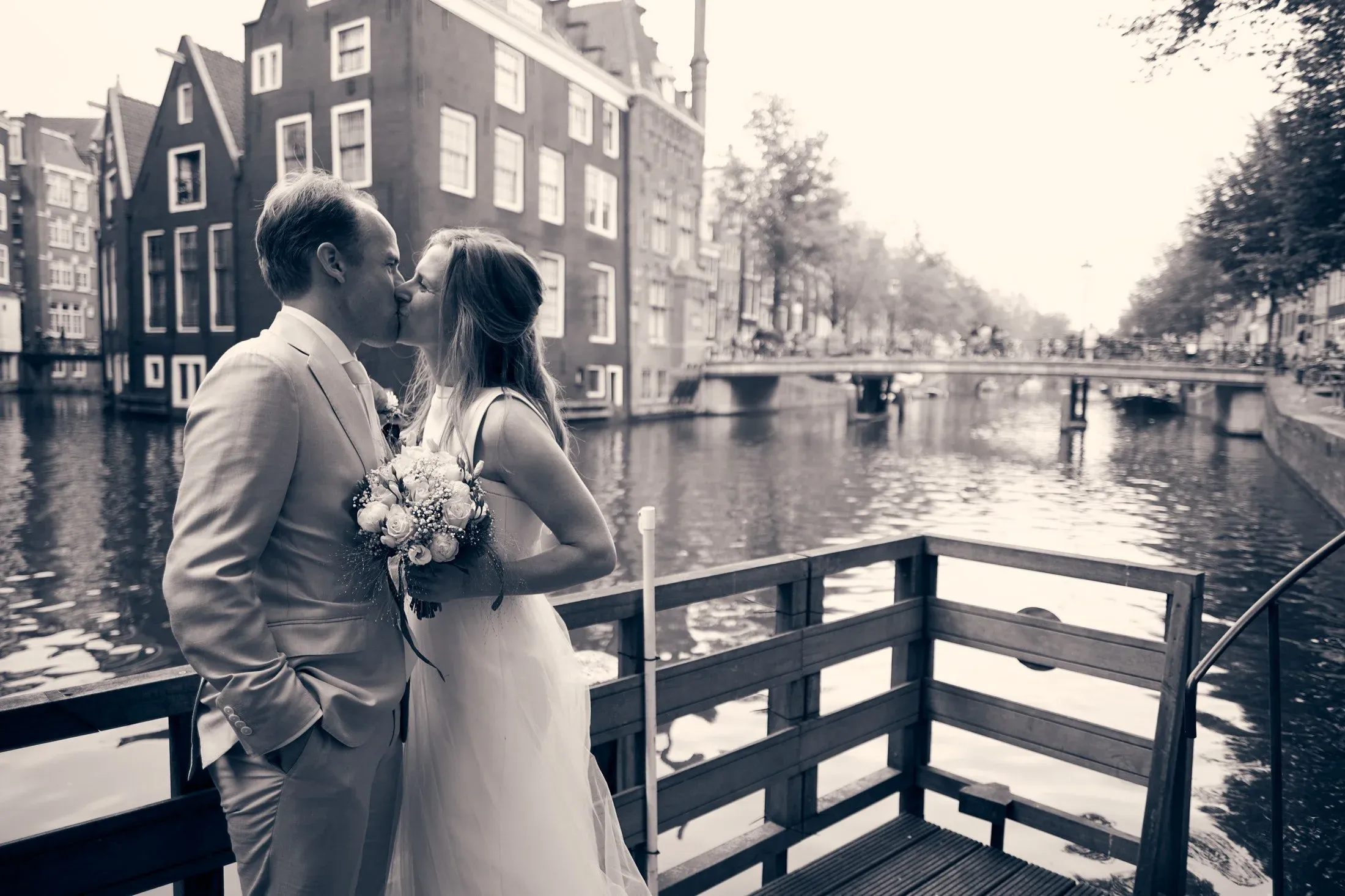 Een getrouwd paar kussen elkaar op een brugje over een gracht in een stad, met historische gebouwen op de achtergrond.
