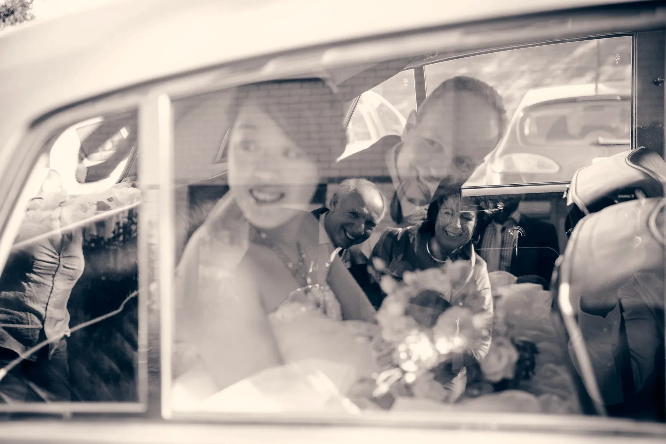 Vrouw en man kijken door een autodeurruit, lachend en poseren voor een foto. Bijsnijdingen van andere mensen en auto's zichtbaar buiten de auto.