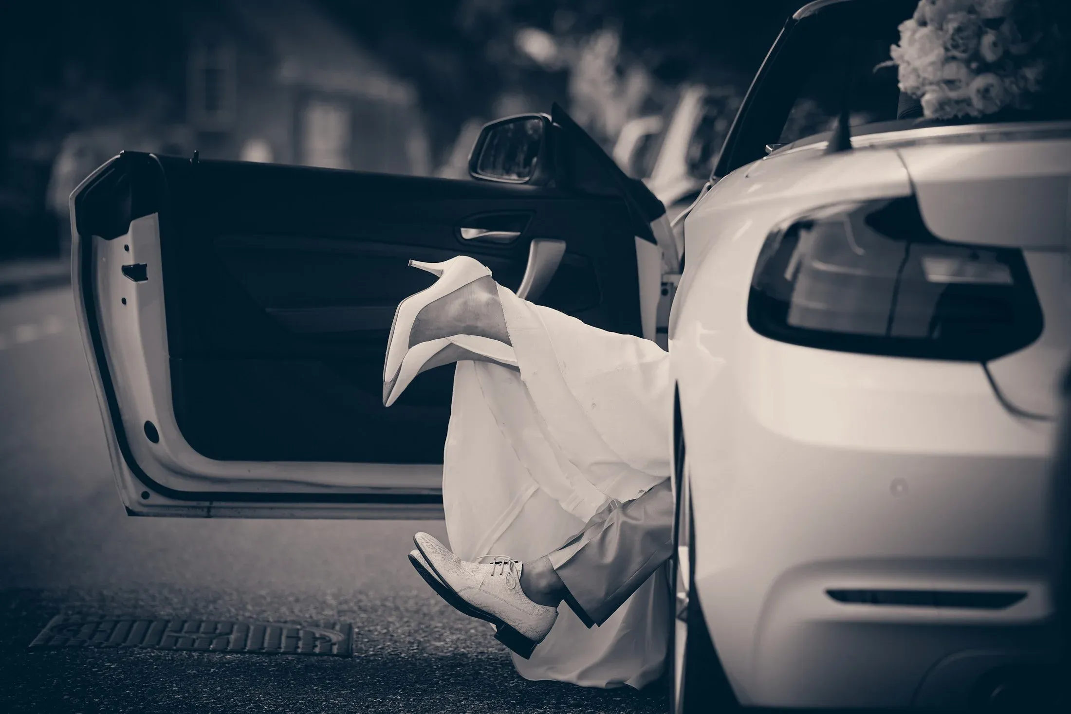 Een persoon met witte schoenen en kleding die uit een witte auto stapt, met benen omhoog op de rand van de auto. Het is zwart-witbeeld op een stadsweg.