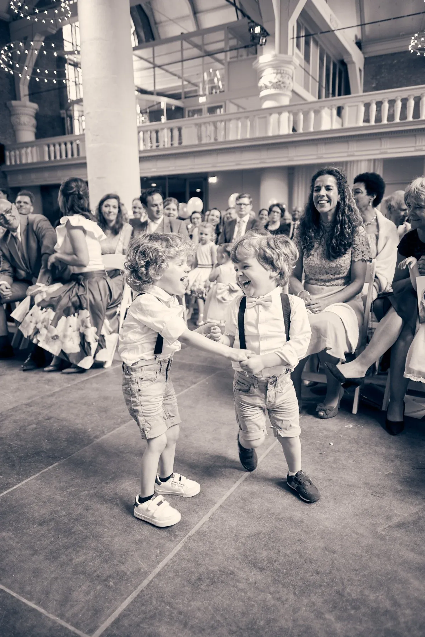 Twee jonge kinderen dansen en lachen samen op een feest, omringd door een lachend publiek.