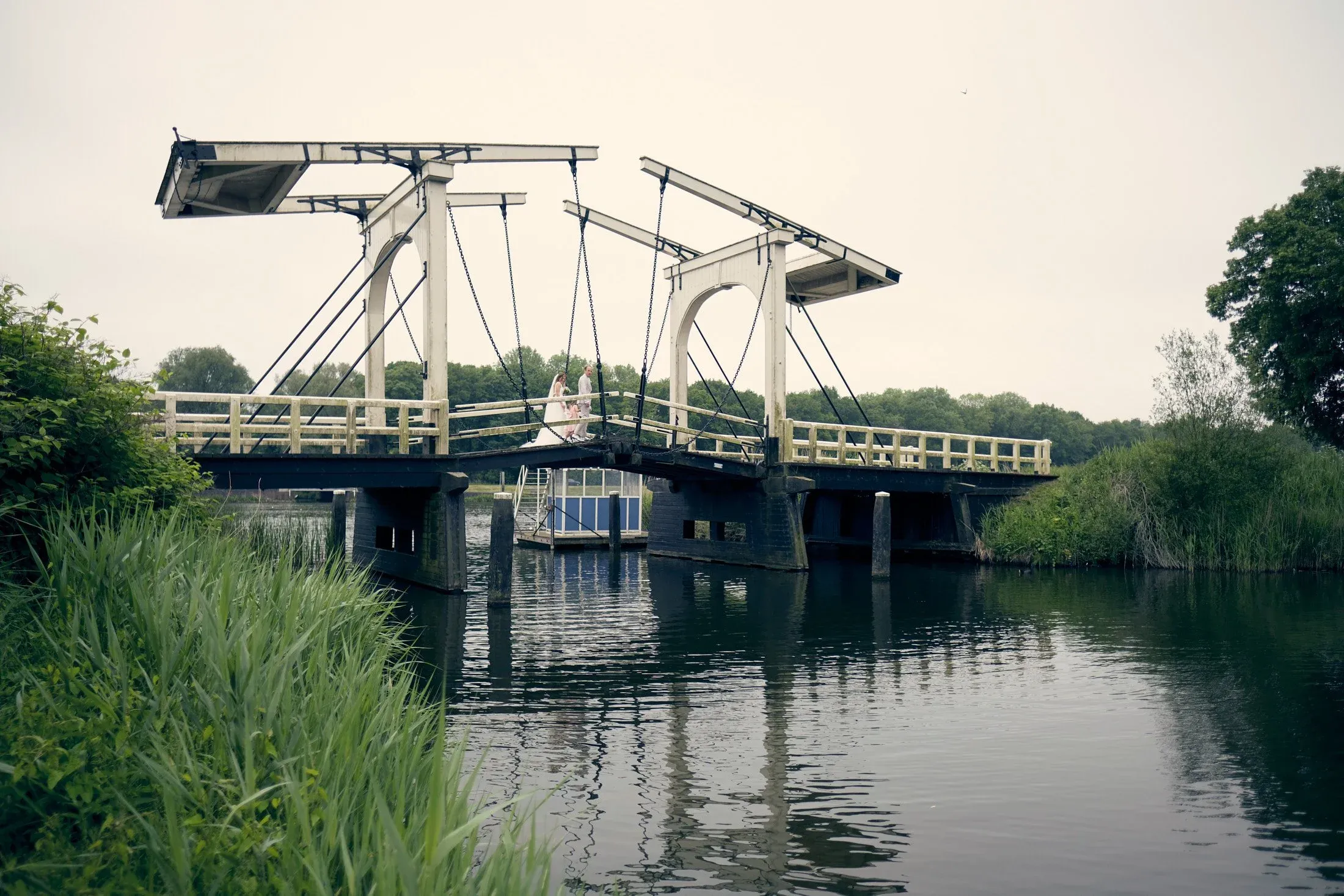 Een houten brug met schommelende delen over een rivier, met drie mensen erop en groene begroeiing aan de zijkanten.
