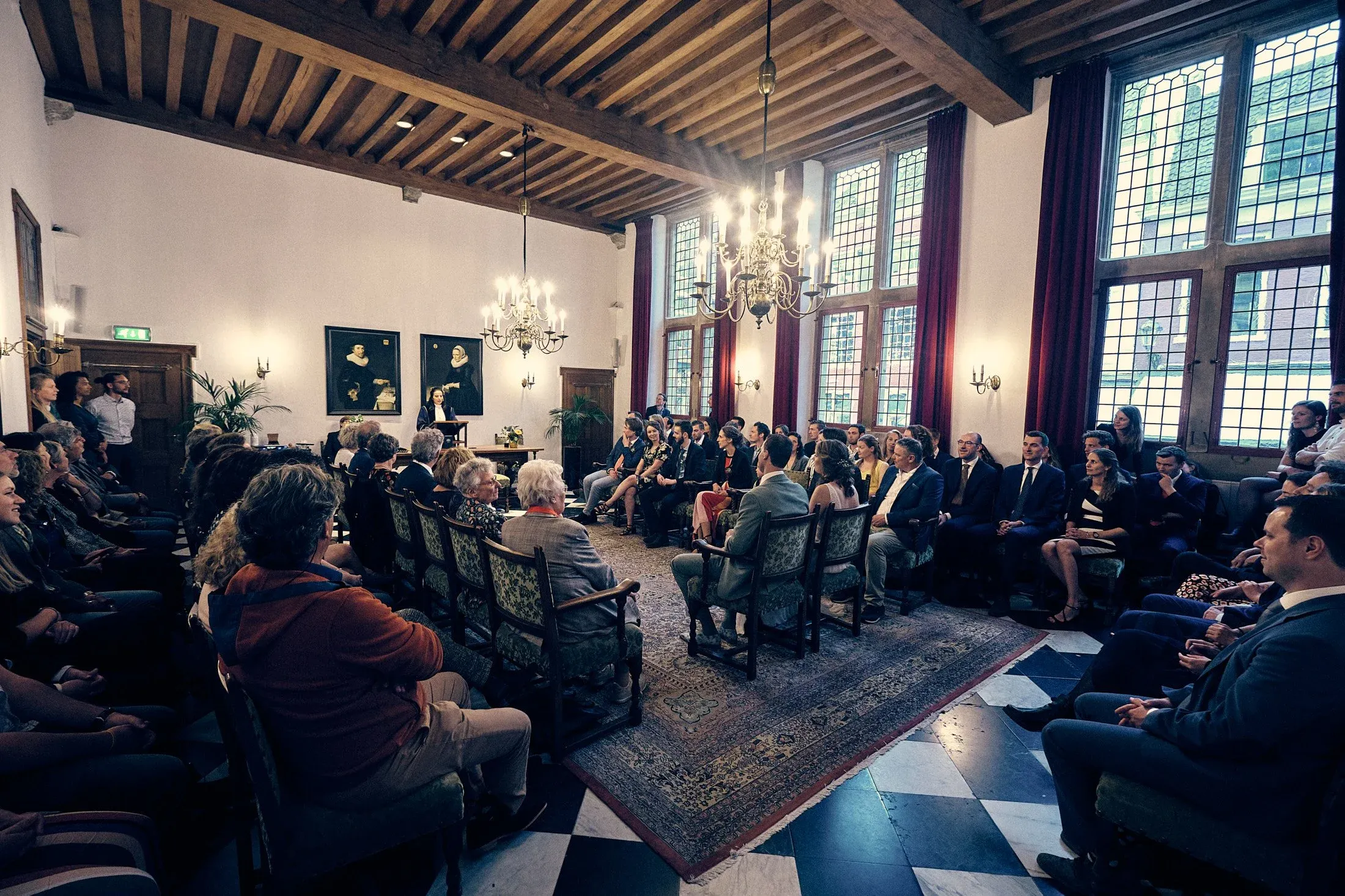 Een grote formele bijeenkomst in een klassieke, elegante zaal met hoge ramen, grote kroonluchters en houten plafond, waar mensen luisterend naar een spreker zitten.
