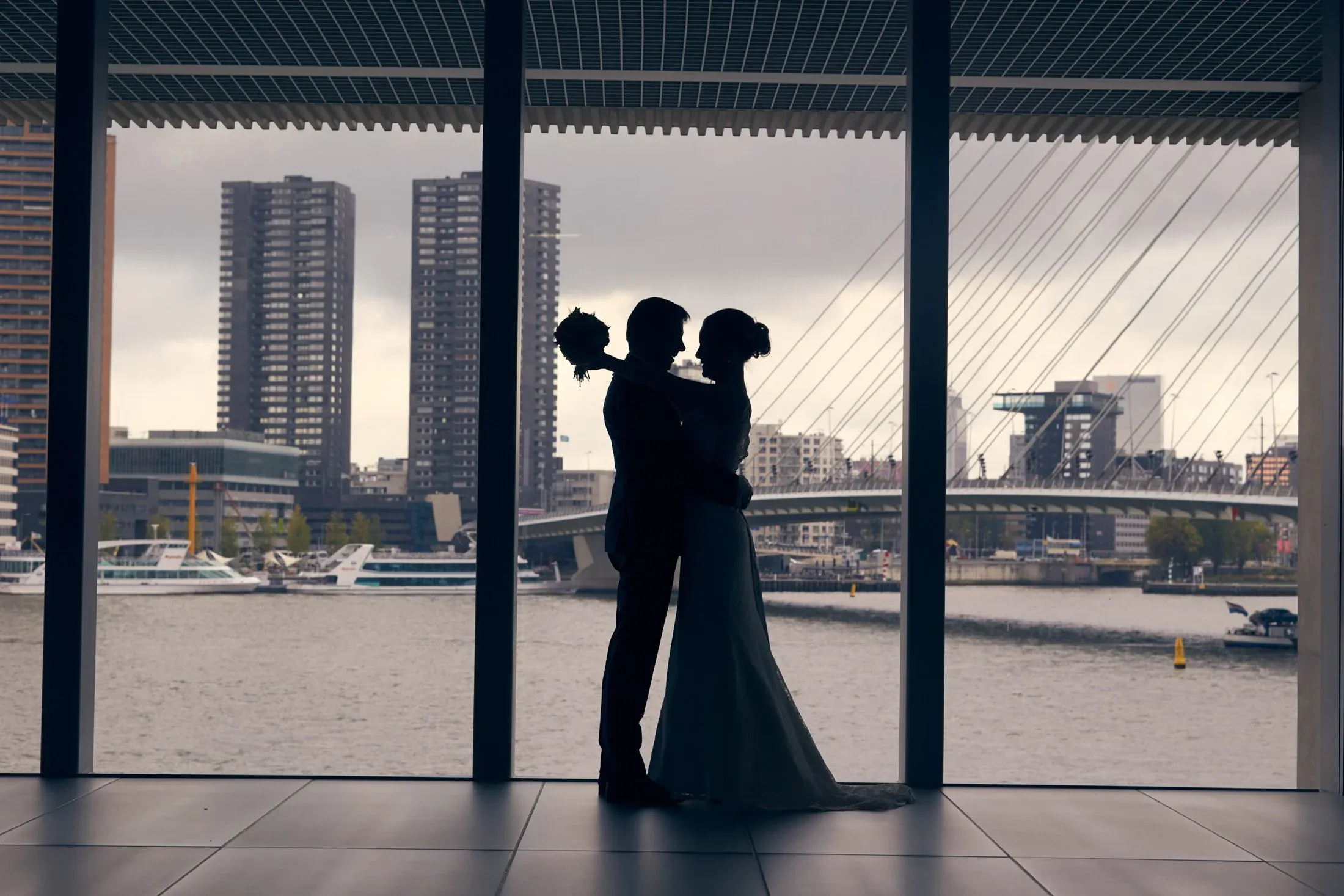 Silhouet van een bruidspaar voor een groot raam met uitzicht op de Erasmusbrug