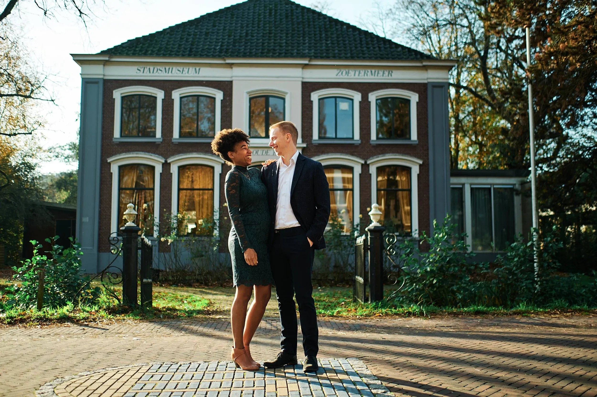 Een lachend paar staat samen voor een historisch stadsmuseumgebouw, omgeven door bomen, in de herfstlicht.