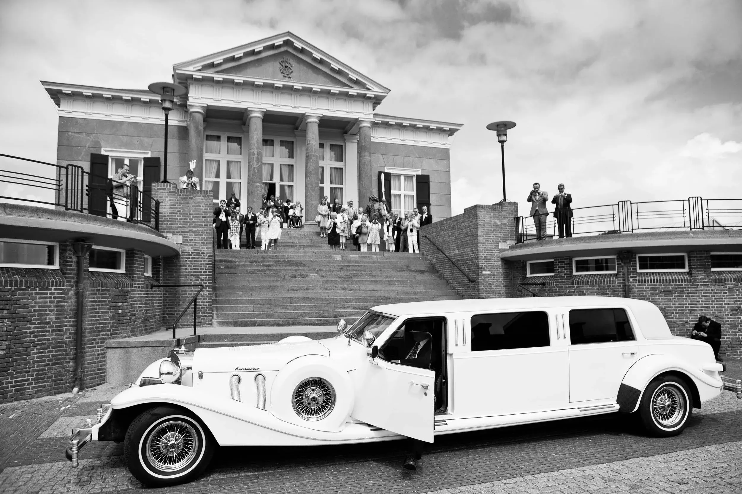 Een zwarte en witte foto van een bruiloft met een oude limousine voor een groot gebouw, mogelijk een kerk. Mensen staan op de trap en langs de rand, sommigen nemen foto's, anderen praten of kijken toe. De limousine staat op de weg voor het gebouw.