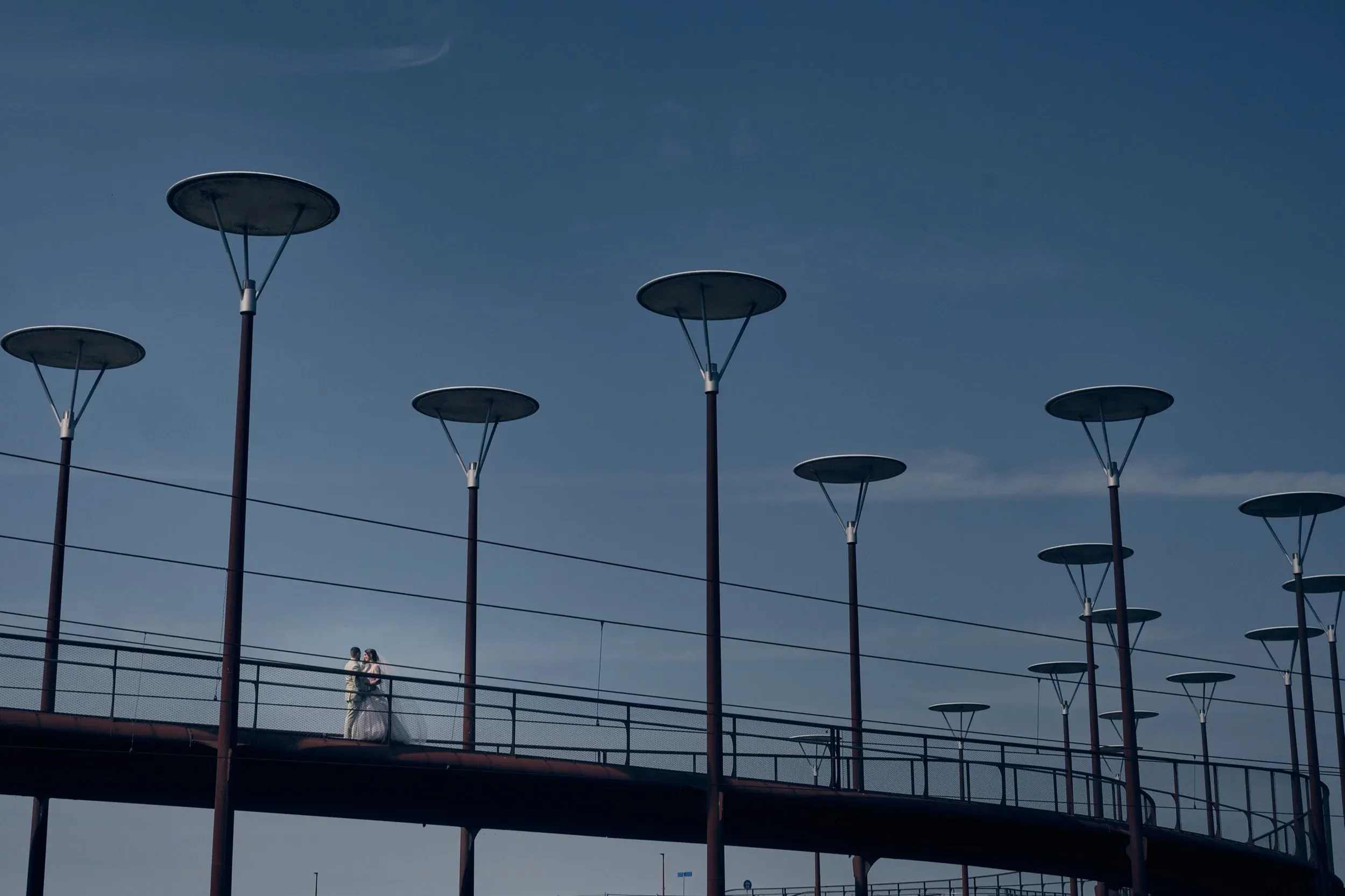 Een bruid en bruidegom staan samen op een brug, omgeven door straatlantaarns tegen een blauwe hemel.