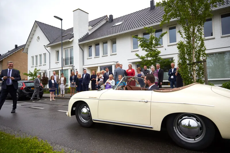 De witte Porsche rijdt de straat in, de gasten staan te wachten