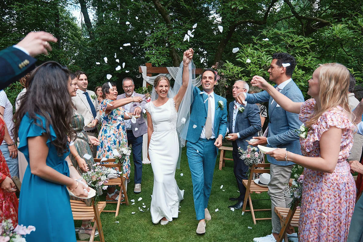 Confetti dwarrelt over Karien en Niels terwijl ze juichend tussen de gasten door lopen