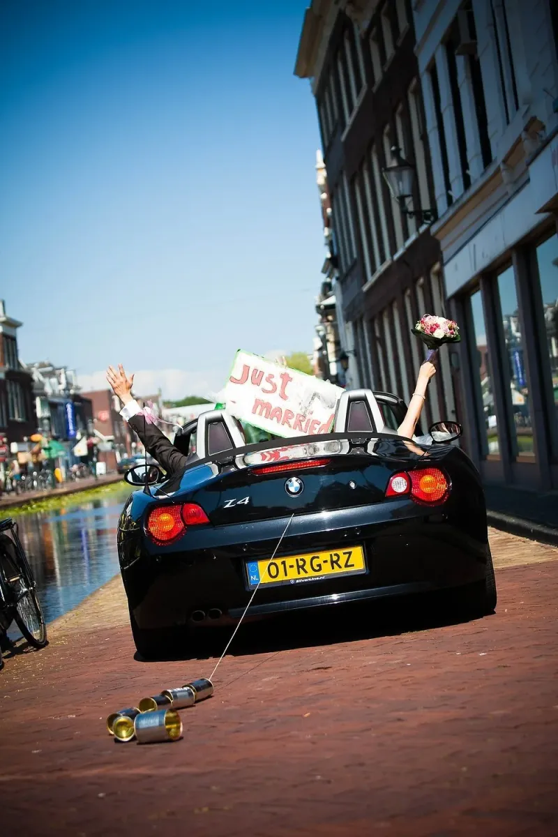 Zwarte BMW Z4 cabrio met ‘Just Married’-bord en blikjes, het bruidspaar zwaait uit de open auto