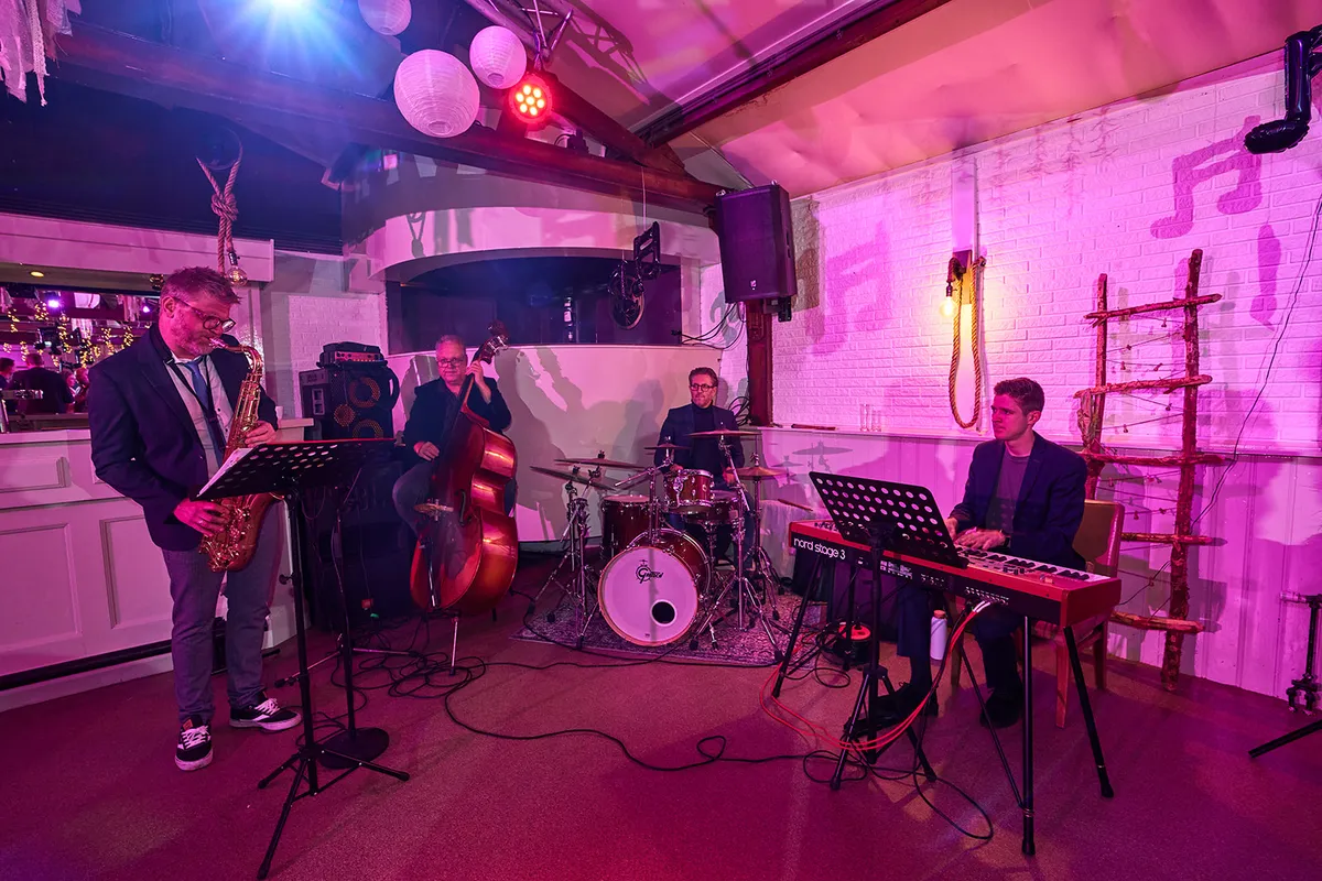 De liveband speelt in roze licht, met saxofoon, contrabas, drums en keyboard