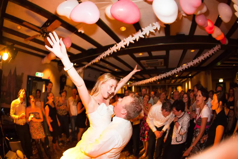 De bruidegom tilt de bruid op de dansvloer op, omringd door klappende gasten onder roze ballonnen