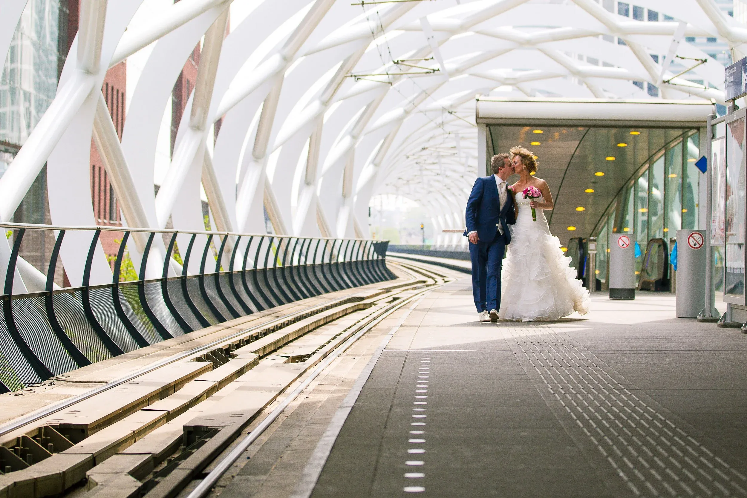 Twee mensen in bruiloftskleding lopen samen in een moderne stationshal, de man in een blauw pak en de vrouw in een witte trouwjurk, ze kussen elkaar.