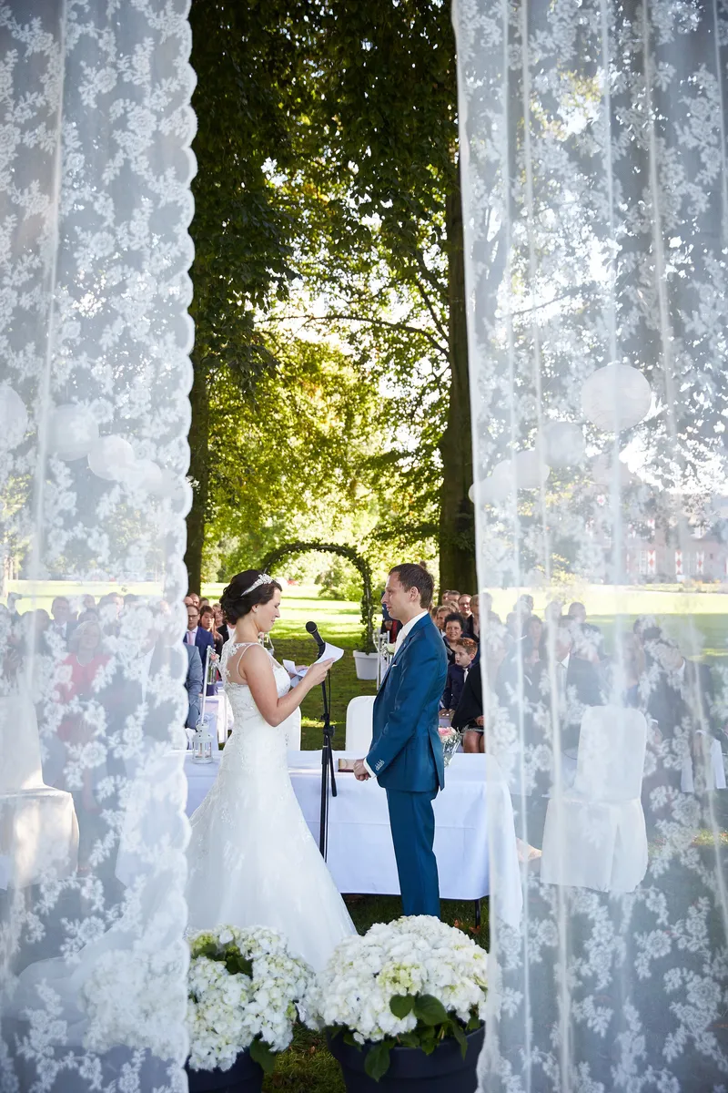 De ceremonie in de tuin, gezien door een kanten gordijn