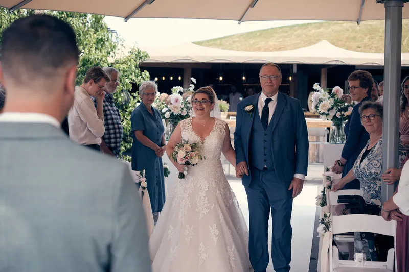 Angelique loopt met haar vader over de witte loper tussen de gasten