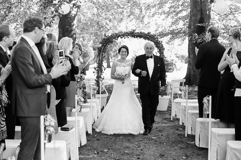 Frederike loopt met haar vader onder de boog door naar de ceremonie, in zwart-wit