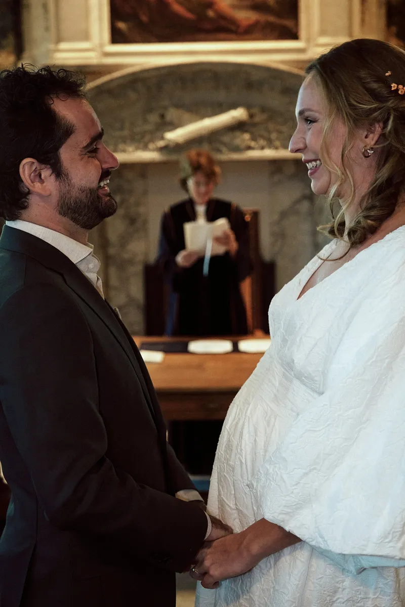 Anouk kijkt naar Raphaël, haar buik zichtbaar in de witte jurk