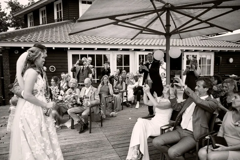 Het bruidspaar loopt over een terras terwijl gasten op stoelen hun telefoons omhoog houden