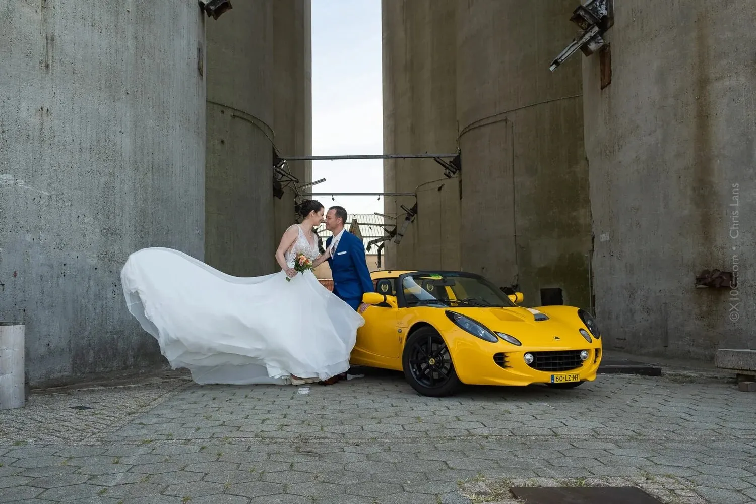 Bruidspaar bij een gele sportauto tussen hoge betonnen silo’s