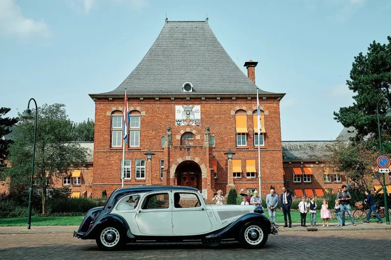 Crèmekleurige Citroën met donker dak voor een bakstenen gemeentehuis met gasten op de stoep