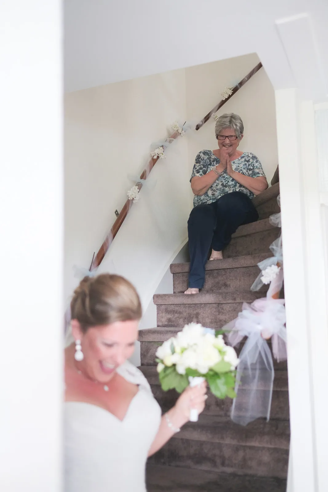 Moeder zit trots op de trap met de handen gevouwen terwijl de bruid lachend naar beneden komt