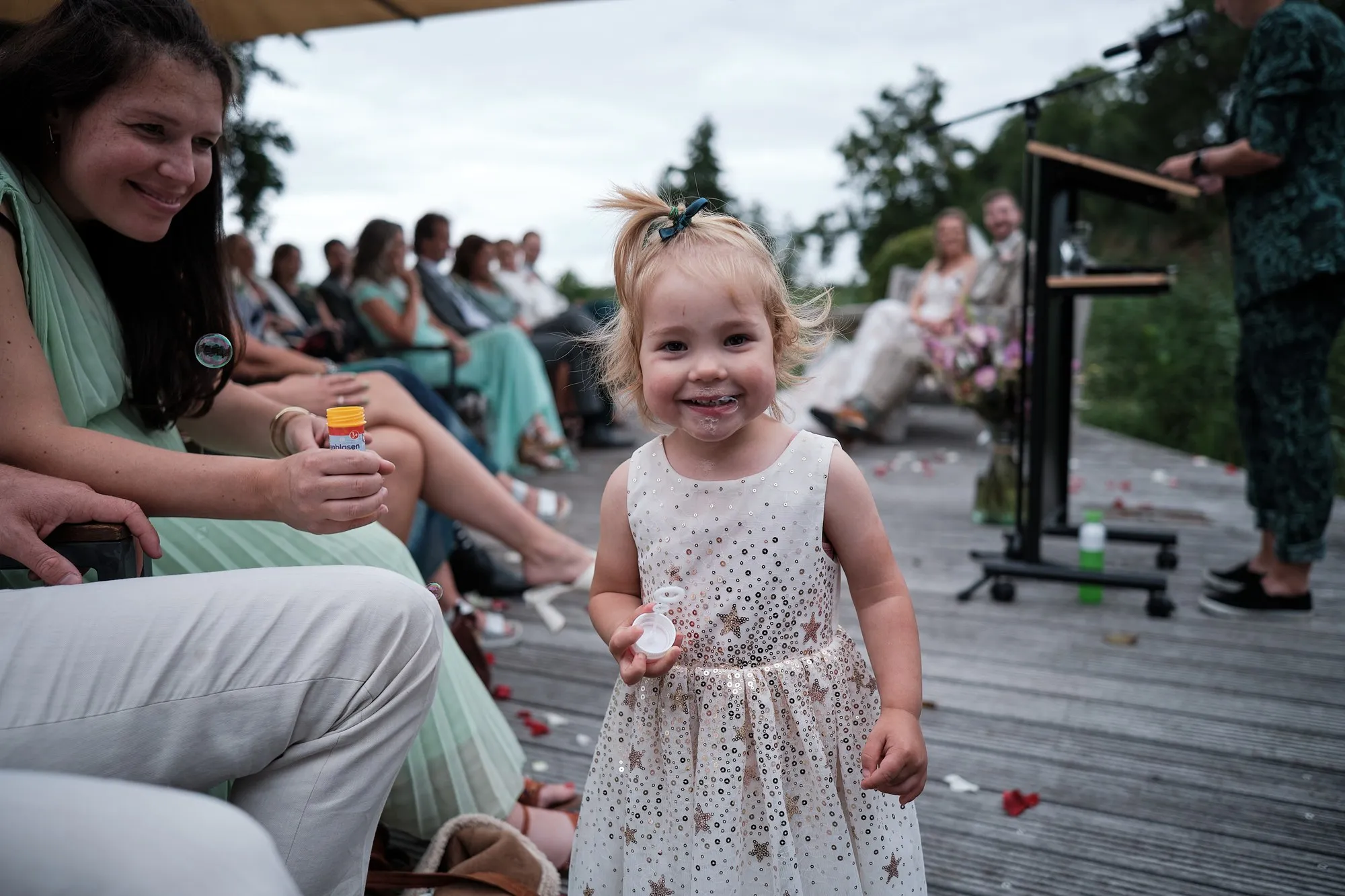 Lachend meisje met een bellenblaas loopt langs de gasten bij een buitenceremonie, het bruidspaar zit op de achtergrond