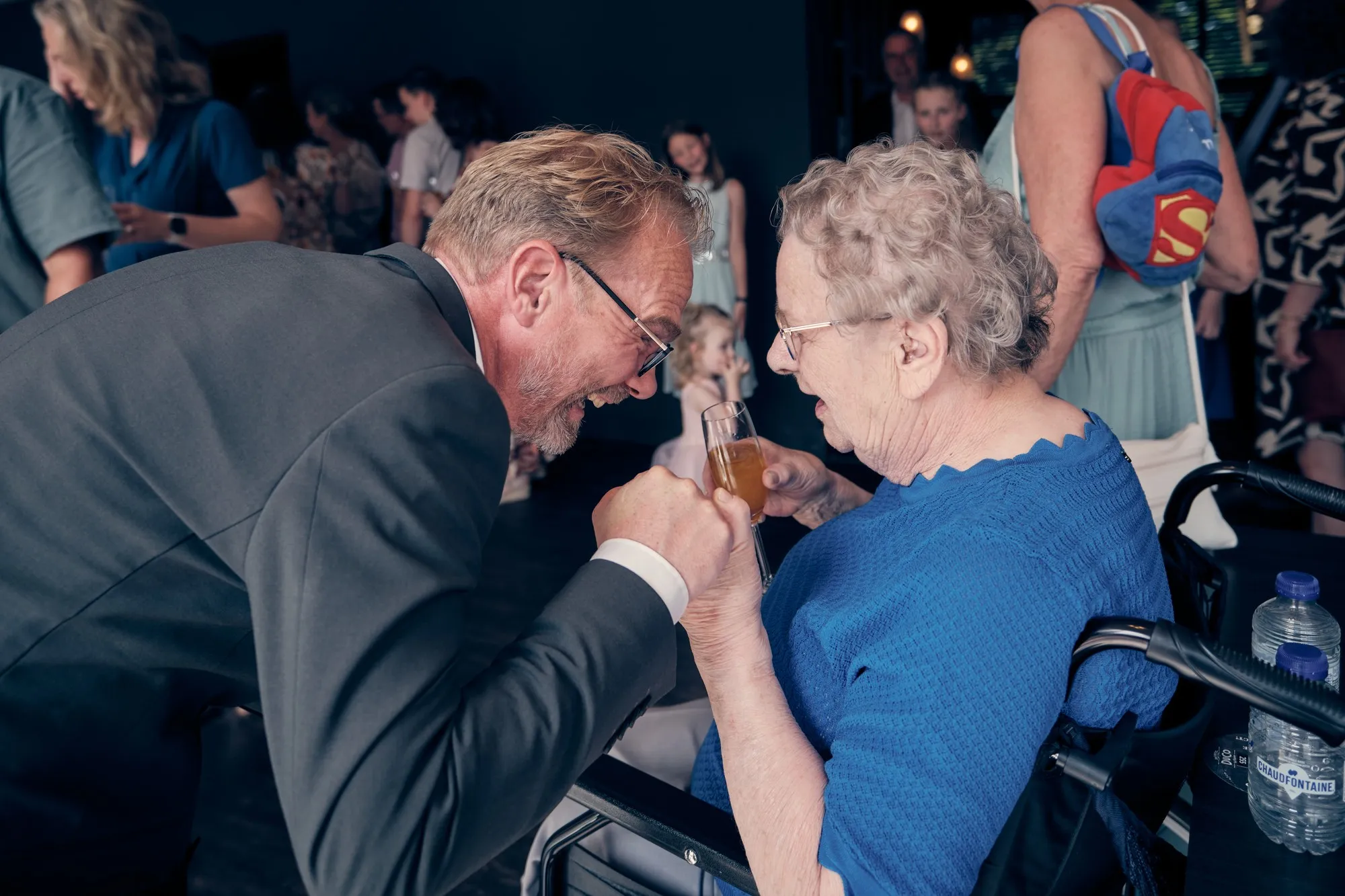 Gast buigt zich lachend naar een oudere dame in een rolstoel en proost met haar