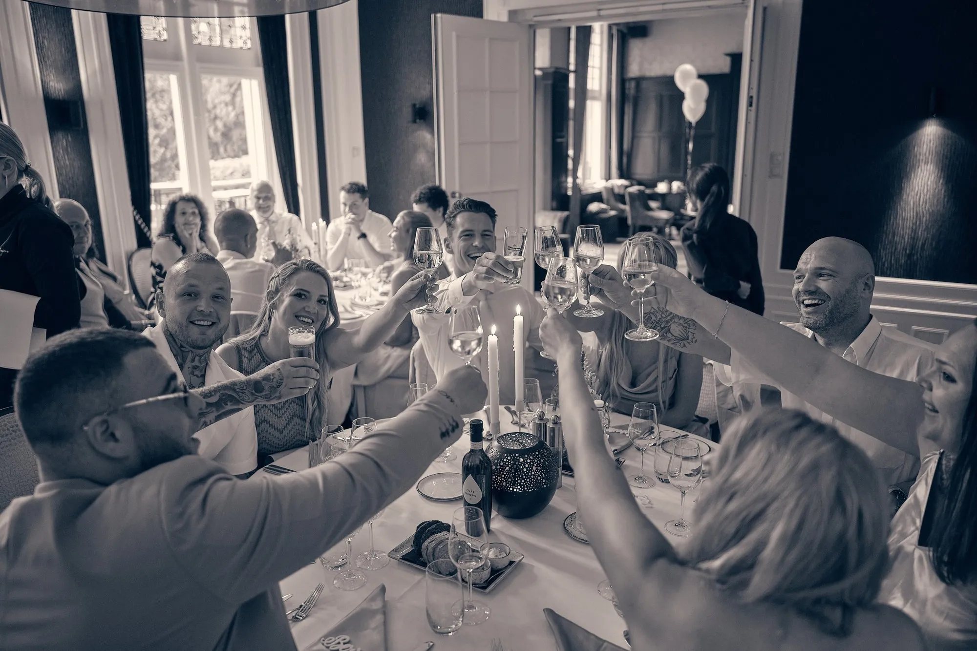 Gasten aan een lange dinertafel heffen lachend het glas, zwart-wit