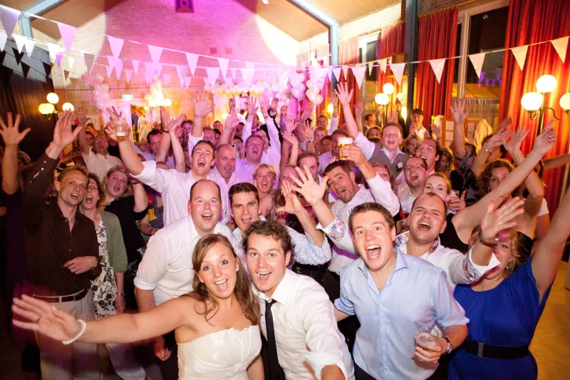 Het bruidspaar springt vooraan op de dansvloer met de feestgroep, handen omhoog onder vlaggetjes en roze licht