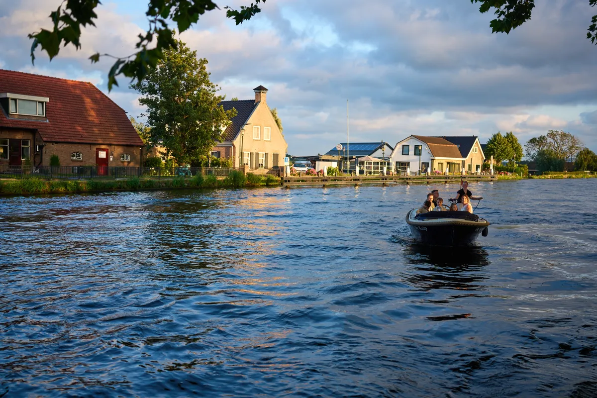 Het bootje vaart over de Vliet in het avondlicht, met de huizen aan de overkant