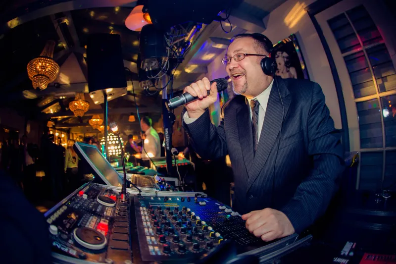 De dj zingt lachend in de microfoon achter zijn mengpaneel, onder kristallen kroonluchters