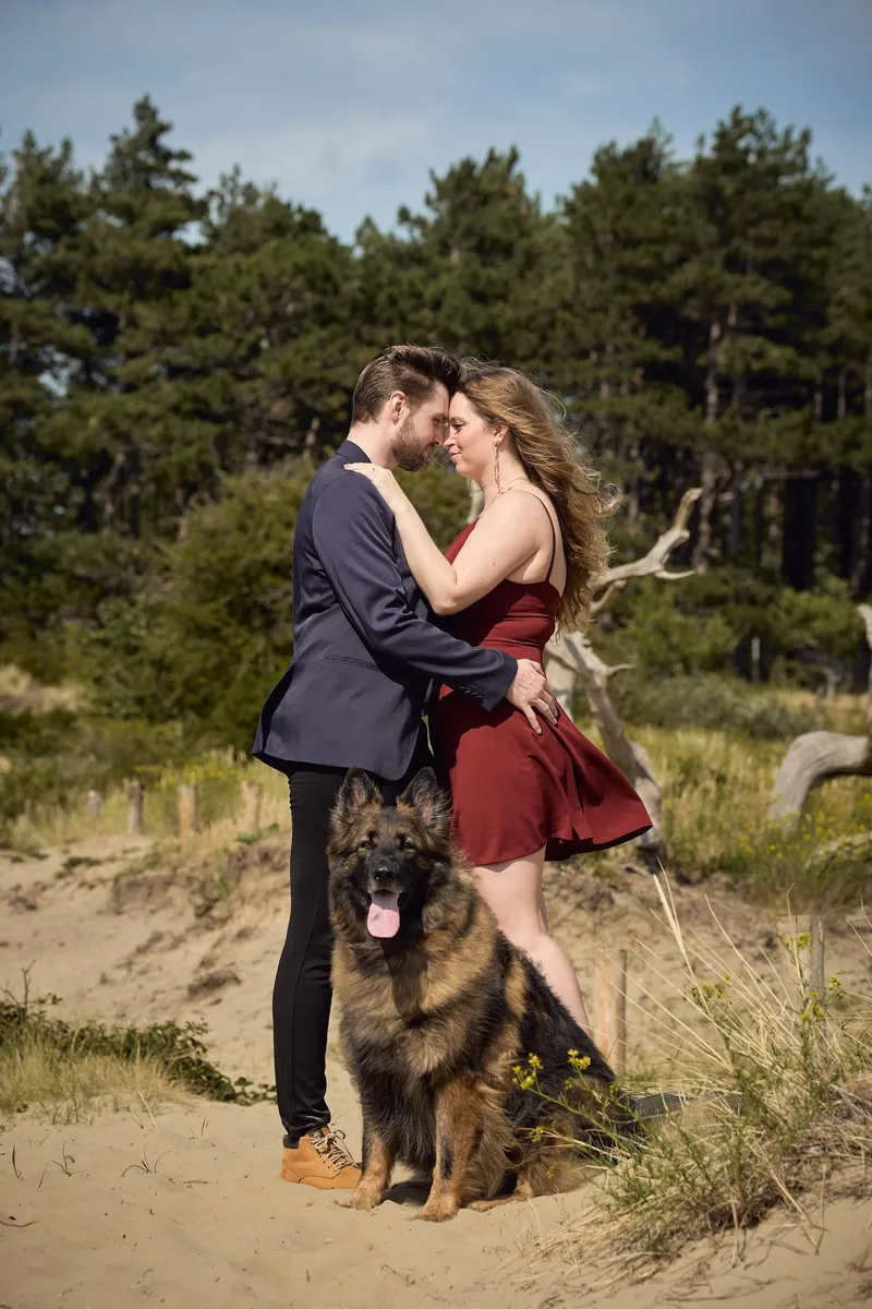 Een koppel kust in de duinen terwijl hun herdershond naast hen zit