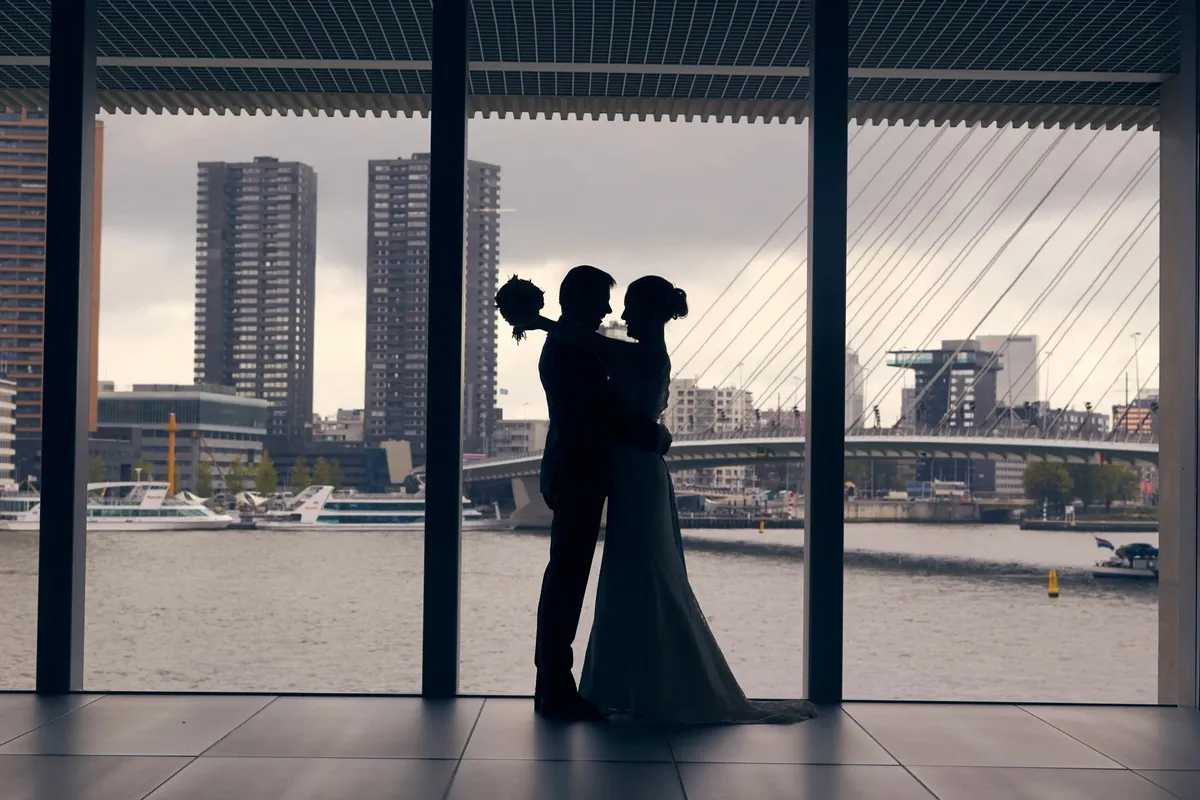 Silhouet van het bruidspaar voor een groot raam in De Rotterdam, met de Erasmusbrug en de torens van de Kop van Zuid