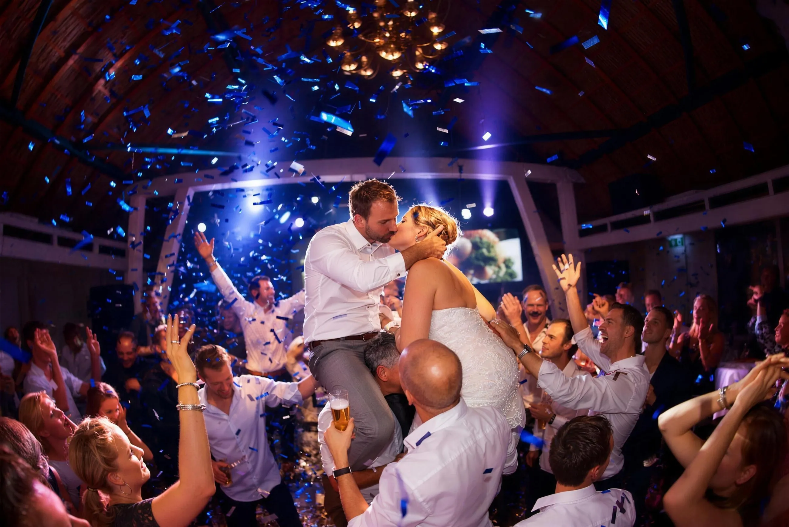 Bruidspaar wordt op de schouders gedragen en kust onder een regen van blauwe confetti