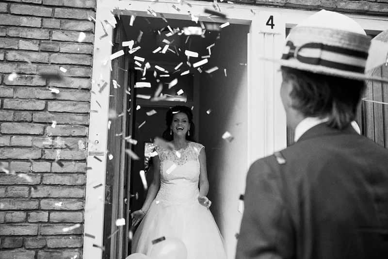 Zwart-wit: de bruid komt lachend door een deur vol confetti, de bruidegom kijkt toe