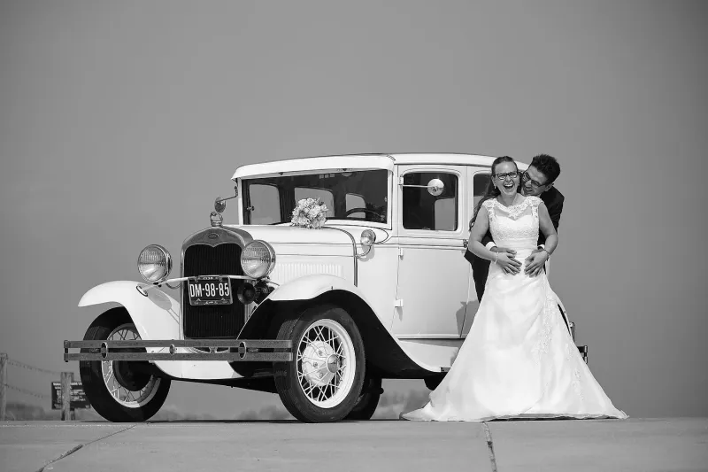 Zwart-wit: een lachende bruid en de bruidegom die haar van achteren omhelst bij een Ford uit 1930