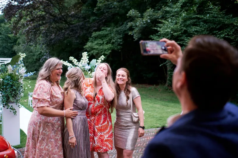 Vier vrouwen lachen in de tuin terwijl een gast op de voorgrond het moment filmt met zijn telefoon