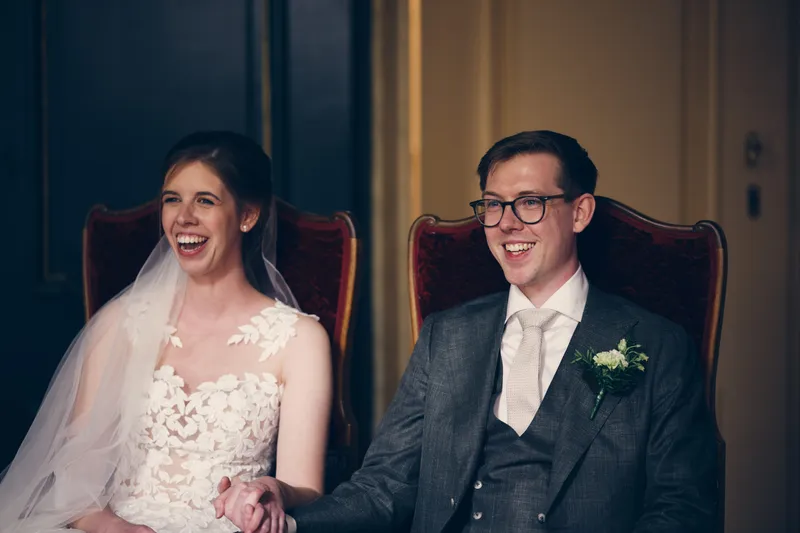 Yvo en Marjolijn lachen naar elkaar op de rode stoelen in de trouwzaal