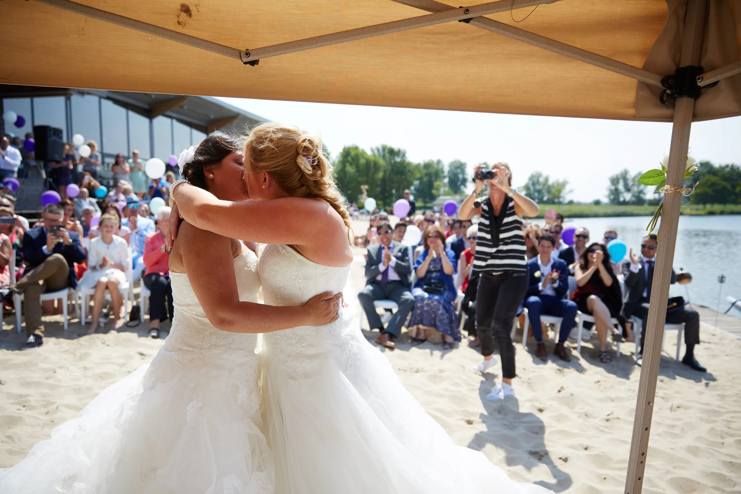 Prunella en Cora omhelzen elkaar onder de parasol op het strand van Villa Westend, met de gasten erachter