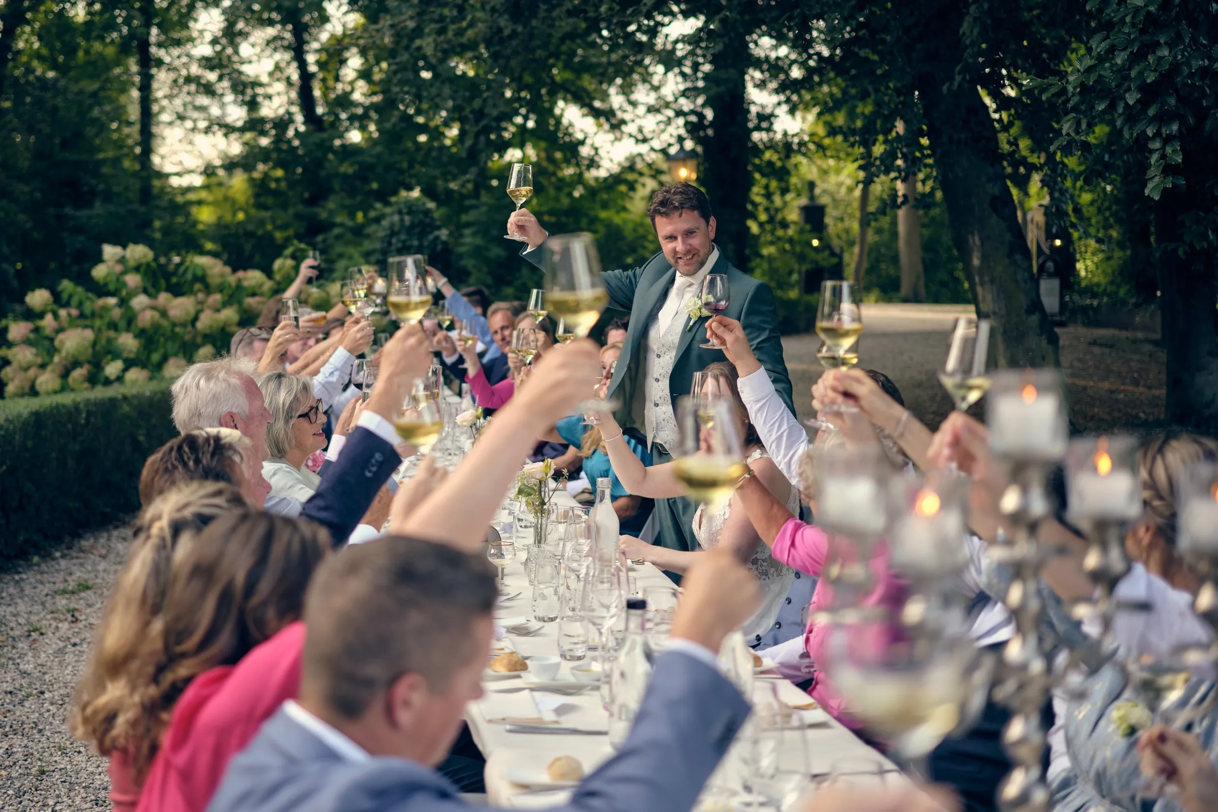 Bruidegom staat op de bank en proost met de gasten aan een lange tafel in de tuin
