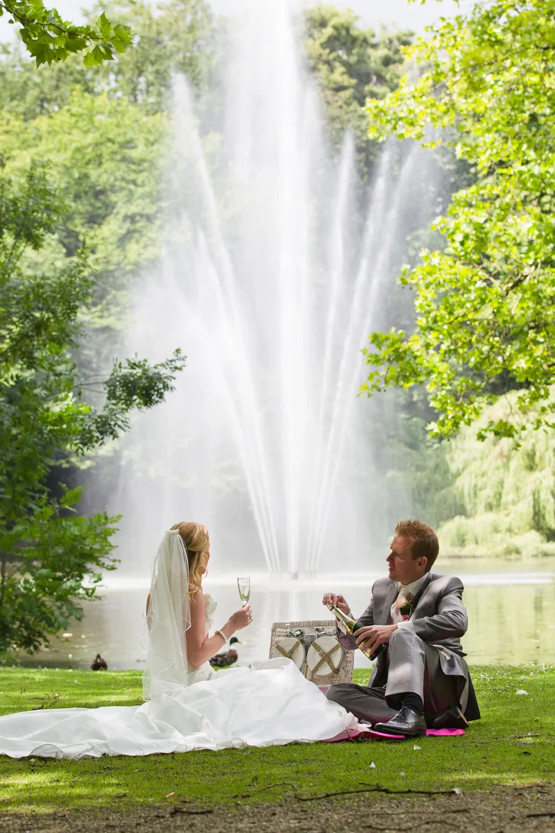 Loes en Jan zitten met champagne in het gras, met de fontein in het Julianapark achter hen