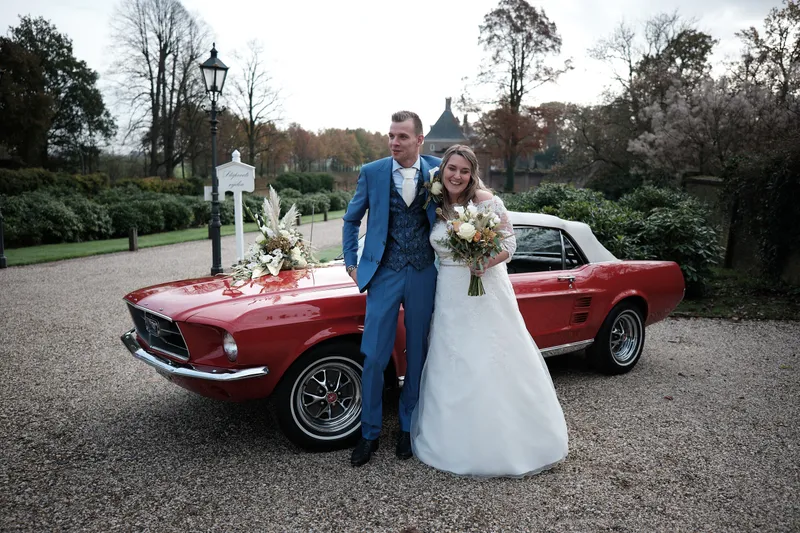 Wendy en Gerrold naast de Mustang, met het poortgebouw erachter