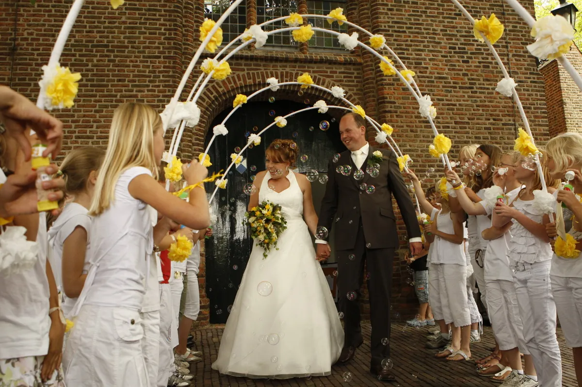 Yvonne en Robert-Jan lopen de kerk uit onder bogen met gele bloemen, gedragen door de kinderen van haar klas