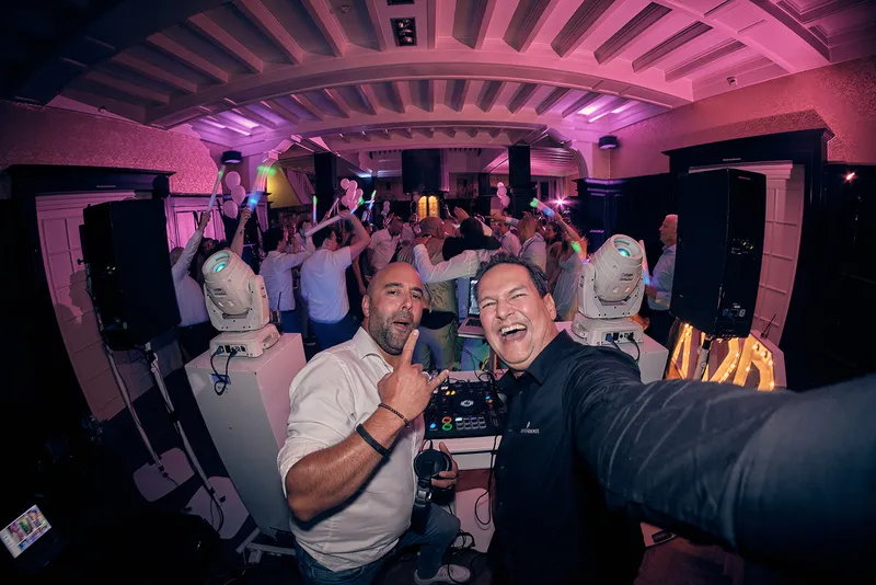 De dj maakt achter de draaitafels een selfie met een gast, de zaal erachter