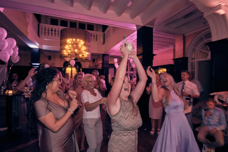 Een gast juicht met het gevangen boeket hoog boven haar hoofd, de vrouwen om haar heen lachen en klappen