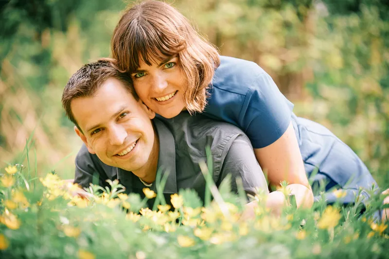 Een koppel ligt lachend in het gras tussen gele bloemen, zij in een blauwe blouse