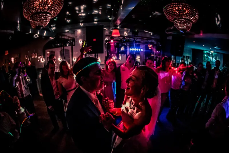 Het bruidspaar danst in een donkere zaal onder rode kroonluchters, de bruidegom met een lichtgevende hoofdband