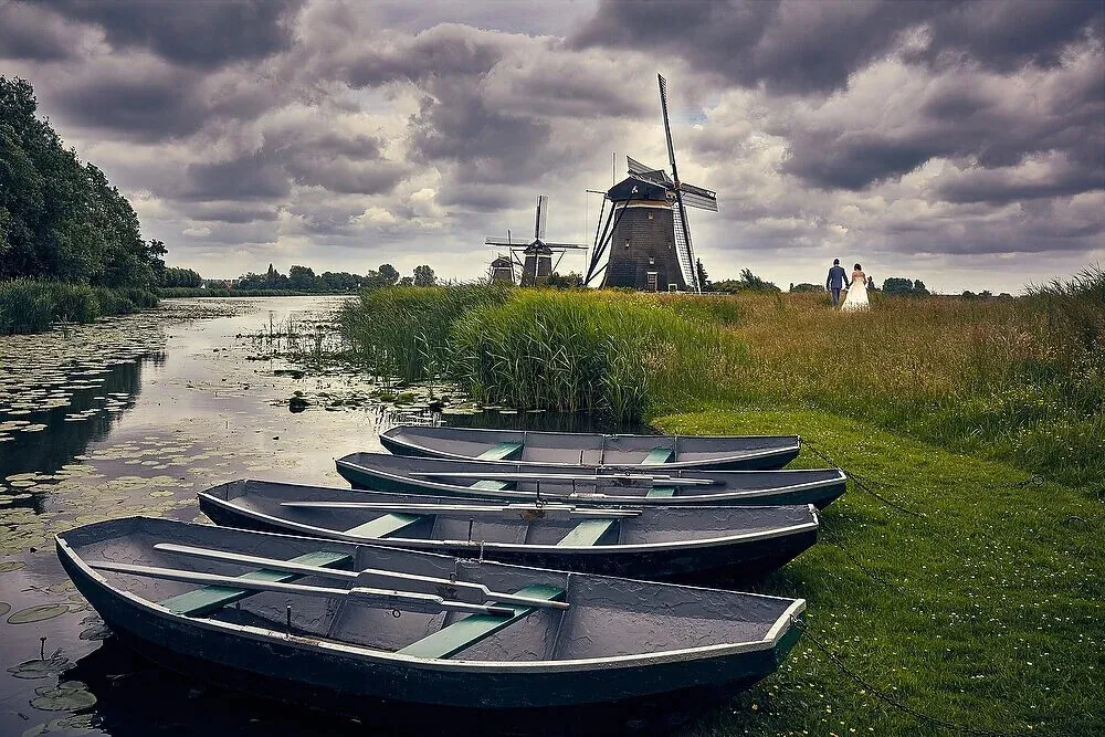 Het bruidspaar wandelt in de verte langs het water bij drie molens, met roeibootjes op de voorgrond